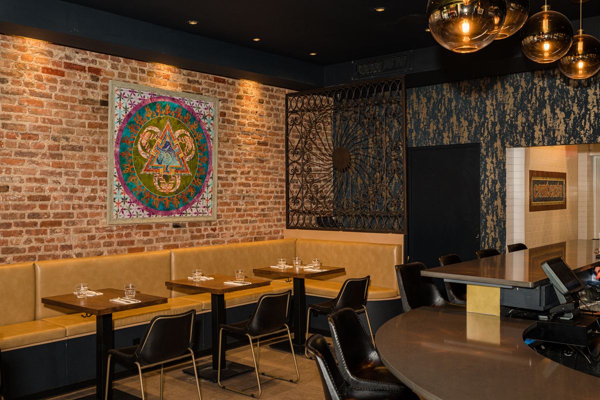  A warm, inviting restaurant space with exposed bricks, mustard half banquettes, paired with dark brown tables and leather chairs. Islamic-inspired art hangs on the wall.