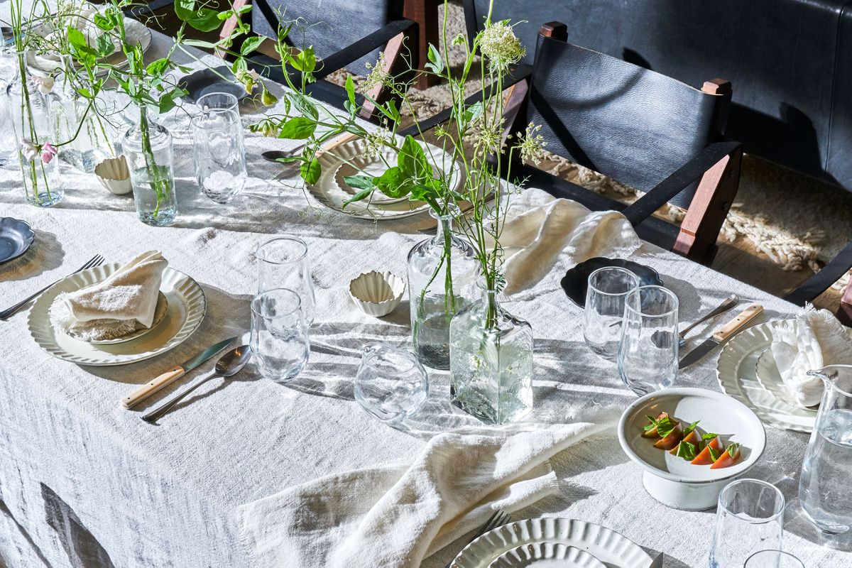  A long dining table covered with a linen tablecloth, place settings, and green floral arrangements in clear glass vases.